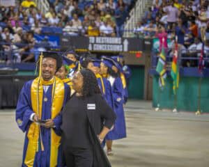 A graduate in cap and gown smiles and walks with a staff member, surrounded by fellow graduates and cheering crowd.