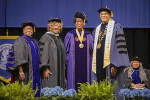 Four people in academic regalia stand on stage, smiling during a graduation ceremony. Blue flowers in foreground.