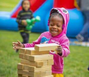 A young girl in a pink hoodie joyfully stacks wooden blocks outdoors, with a bouncy castle in the background.