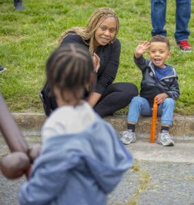 A smiling child waves while sitting next to an adult on grass, with another child in a blue jacket facing away.