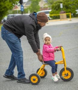 An adult helps a child ride a yellow tricycle on a paved surface, surrounded by greenery.