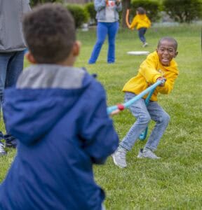 Two children play tug-of-war on grass, with one laughing and pulling a rope while others engage in games nearby.