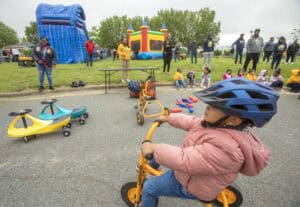 A child in a helmet rides a tricycle at a community event with inflatables and spectators nearby.