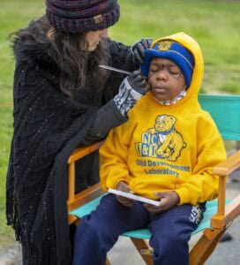 A child in a yellow hoodie sits still while an artist paints designs on their face outdoors.