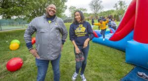 Two smiling adults stand in a colorful outdoor setting with inflatable play equipment and bouncing children nearby.