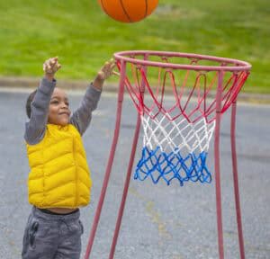 A young boy in a yellow vest joyfully reaches to shoot a basketball into a hoop outdoors.