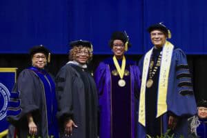 Four individuals in academic regalia pose together at a graduation ceremony, smiling under a blue backdrop.