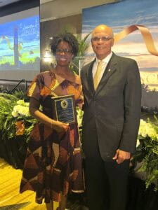 Two individuals are smiling and posing together, holding an award plaque against a decorated backdrop.