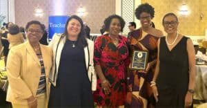 Five women pose together, smiling, at an event, one holding an award, with festive decor in the background.