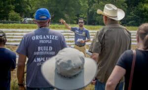 A speaker gestures while discussing farming practices to an attentive audience in a field.