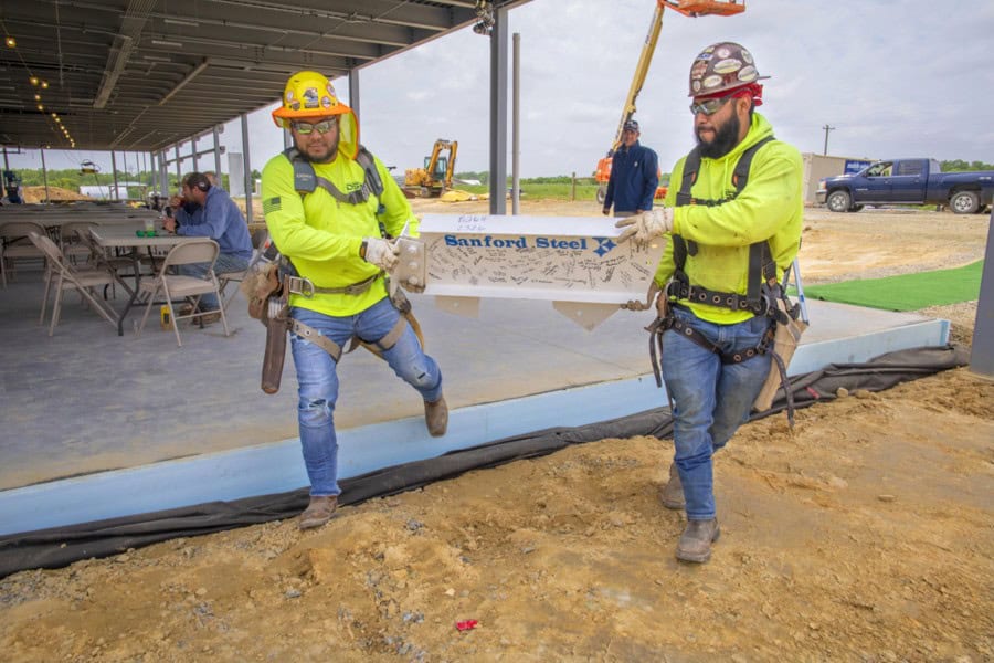 Two construction workers in safety gear carry a steel beam marked "Sanford Steel" at a building site.