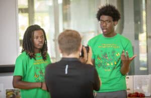 Two young men in green shirts discuss pest management while one is being photographed.
