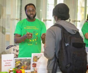 A man in a green shirt speaks to a person with a backpack at an information table about pest management.