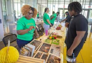 A woman in a green shirt demonstrates gardening practices at a booth displaying fresh produce and informational…