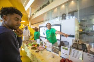 A woman in a green shirt points while discussing bees at a table with food items and informational posters. A young man…