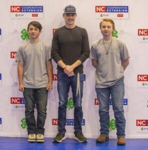 Three young men stand together against a backdrop featuring the NC Cooperative Extension logo.