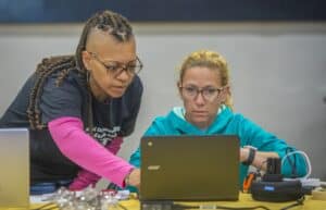 Two women collaborate at a laptop, focused on a task, with technology tools around them.