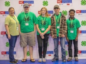 A group of five individuals in green shirts pose in front of a banner for Cabarrus County 4-H.