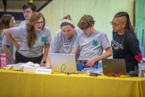 A group of kids and adults collaborate at a table with a laptop and drone, focused on a project.
