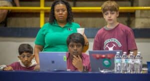 A group of young participants in matching shirts focus on laptops at a drone competition, with a mentor guiding them.