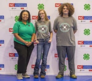 Three individuals pose together in front of a backdrop, representing NC Cooperative Extension with logos and a green…