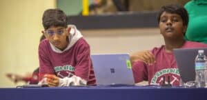 Two boys at a table with laptops, focused on a task. One wears glasses and a hoodie, the other gestures while thinking.