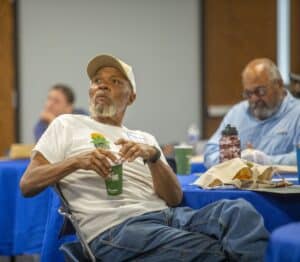 A man with a gray beard and beige cap sitting at a conference table with a green cup in hand.