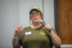 A woman speaking into a microphone at a conference or seminar, wearing a green cap and T-shirt with.