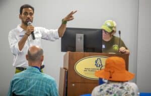 Engaged speaker presenting at conference with audience and technical staff in background.