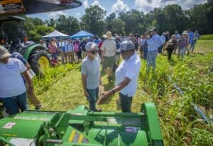 Farmers discuss equipment at a field demonstration with a crowd observing in the background.