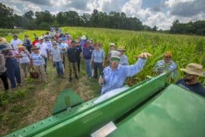 A diverse group of people gathers around a farm machinery demonstration in a cornfield under a partly cloudy sky.