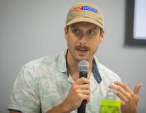 Man speaking into microphone at CAES News event wearing a beige cap and casual shirt.