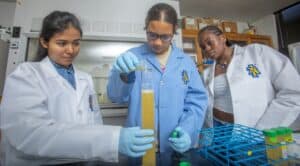 Three students in lab coats conduct an experiment, pouring liquid into a measuring cylinder in a laboratory setting.