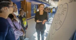 A person in a blazer presents to a group of students near a poster during an event in a school hallway.