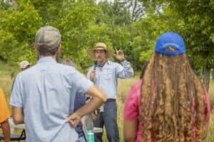 A man giving a speech outdoors to a small group of people in a green, natural setting.