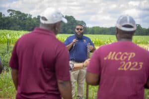 Agricultural expert giving a presentation outdoors to two farmers in a green field.