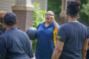 A coach enthusiastically guides a group during an outdoor fitness session, holding a medicine ball.