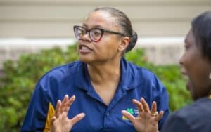 A woman in a navy polo gestures animatedly while speaking, with a blurred figure beside her. Green foliage in the…