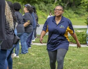 A smiling woman in a navy and yellow shirt walks joyfully in a green park with others in matching outfits behind her.