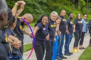 A diverse group of people hold hands outdoors, smiling and engaging with a hula hoop during a community event.