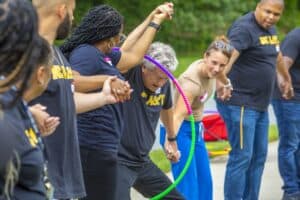 A diverse group of people is holding hands and playing with hula hoops in a lively outdoor setting.
