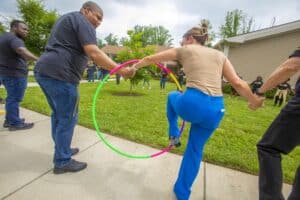 Participants hold hands while stepping through a colorful hula hoop in a grassy area during an outdoor event.