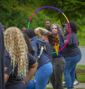 A group of people participates in a playful activity with hula hoops outdoors. Lush greenery surrounds them.