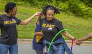 Two women in NC A&T t-shirts participate in a group activity, holding hands and smiling, with a hula hoop nearby.