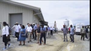A group of people touring an industrial facility or farm, with a large building and storage tanks in.