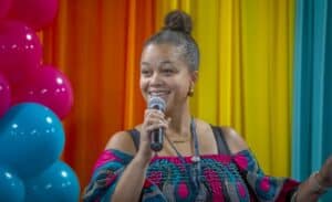 A woman with a microphone smiles while speaking, dressed in a colorful patterned top, in front of vibrant backdrops.