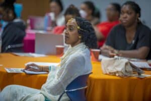 A woman with curly hair, adorned with jewelry, listens attentively at a gathering with others in the background.