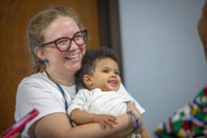 A smiling woman holds a baby in her arms, both looking joyful in a warm setting.