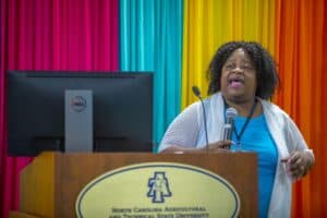 A speaker passionately addresses an audience at a conference, with colorful drapes in the background.