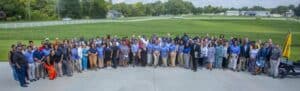 Large group of people in coordinated attire, gathered outdoors on a sunny day with green fields in the background.